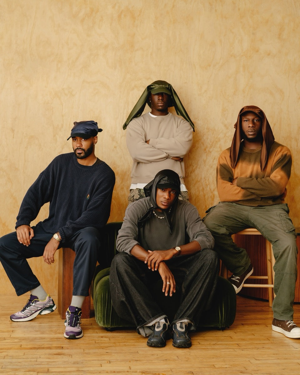 Four men sitting on stools against a beige wall.